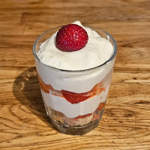 A glass filled with parfait, a dessert consisting of a layer of crushed biscuits, chopped strawberries in syrup, and a cream made made of whipped cream, yoghurt and mascarpone. The dessert has a whole strawberry on top